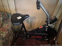 Side profile of the Roadmaster exercise bike showing black padded seat, frame, and weighted flywheel cover with branding.