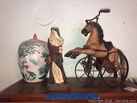Photo showing the ceramic jar, carved wooden Santa Claus figurine, and vintage-style metal horse tricycle figurine together on shelf.
