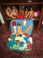 Full view of assorted dolls arranged on a shelf and on the floor in front of a blue suitcase with clothing