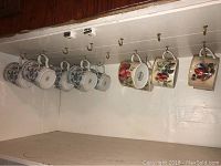 Eight decorative mugs hanging on hooks, displaying various floral and fruit designs.