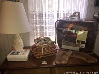 Overall view of lamp, comforter set, first aid kit, baskets, pinking shears and harmonica on a wooden surface in front of lace curtains