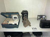Wide shot showing blue carrying case with slides, Argus C3 vintage camera with leather telephoto lens case, and Polaroid style camera on white surface