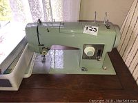 Side view of a green vintage sewing machine with number tag 123 on wooden surface near lace curtained window.