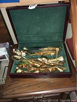 Open wooden case with green velvet lining containing gold-tone forks, spoons, and knives.