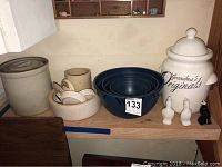 Photo shows a wooden shelf with multiple ceramic kitchen items including stoneware crocks in various sizes, nested blue bowls, a large white labeled crock, and two small chicken-shaped pie vents.