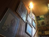 Wall displaying multiple framed artworks of various sizes featuring wildlife, landscapes, sketches, and architectural images under warm lighting on wood paneling.