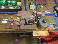 Photo showing hand drill, reddish metal support blocks, metal containers with brass knob tops, small drill bit sets, and packaging on a wooden table.