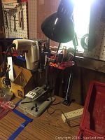Bench-top drill press, black adjustable table lamp, and white power strip on a workbench near a pegboard, showing layout and condition of items.