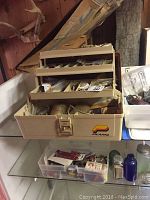 Beige plastic organizer box with multiple tiered trays open showing various leatherworking materials and tools inside.