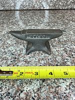 Side view of miniature cast iron anvil beside measuring tape showing length under 6 inches; raised marking visible