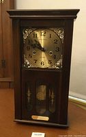 Front view of wooden wall clock showing brass dial, glass door and plaque