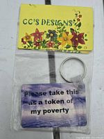 Front of packaged keychain showing "Please take this as a token of my poverty" text on purple background and header card