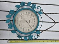Front view of turquoise floral metal outdoor thermometer with chain hanger and ruler for scale