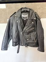 Front view of black leather Brunswick motorcycle jacket hanging; shows overall style, zip pockets, waist belt