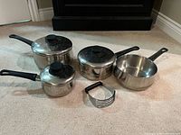 Four stainless steel pans with three lids and pastry blender on carpet