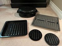 All six pieces laid out: oval roaster with lid, grey perforated pan, black ridged tray, two round trivets