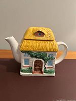 Front view of cottage-shaped ceramic teapot showing door, windows, foliage