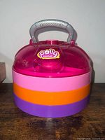 Closed carry-case view showing pink translucent top, stacked color tiers, silver handle, Polly Pocket logo