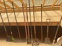 Row of long-handled tools leaning against railing: shovels, rake, rake, post hole digger, shovels