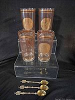 Four amber glass tumblers with gold medallions displayed on clear riser, three ornate spoons laid in front