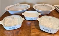 Two square Cornflower Blue casseroles with lids, small open dish, and two larger white casseroles with lids displayed on wooden table