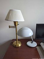 Brass swing-arm lamp with pleated shade next to white dome desk lamp, both on tabletop
