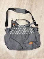 Front view showing gray fabric, white arrow panel, front pockets, brown logo patch, and shoulder strap