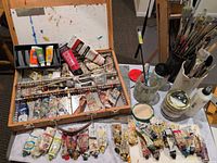 Open wooden sketchbox with numerous oil paint tubes, brushes in containers, mediums on table