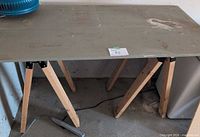 Makeshift workbench with board top on wooden sawhorses; blue fan partly visible