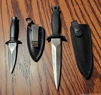 Both stainless steel knives side by side with their matching black leather sheaths on wood surface