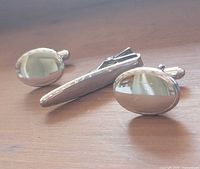 Front view of pair of oval cufflinks and tie clip on wood surface