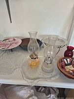 Group shot of two oil lamps, painted wooden bowl, red vase, pink leaf platter, clear platter edge, blue dish, frosted plate on stand
