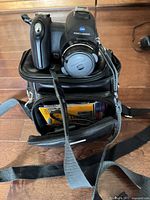 Front view of camera on carrying bag showing lens, AS logo, strap, bag pocket partially open