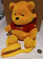 Front view of yellow Winnie the Pooh plush backpack showing red shirt, yellow strap, coin for scale