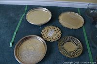 All five serving trays displayed on a blue surface, showing a variety of shapes and designs including a round silver-plated tray with beaded rim and small feet, a round tray with ornate floral engraving and a beaded edge, a round perforated metal tray with lattice design, a decorative round metal trivet, and a silver-plated scalloped edge tray.