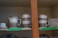 Shelf showing Noritake Progression creamer pitch and 8 stacked teacups with the brown and gold geometric floral band decoration on white porcelain.