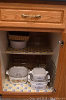 Photo shows open kitchen cabinet with three CorningWare casseroles (two blue cornflower and one floral), a woven basket, clear Pyrex casserole dishes, and pie plates placed on two shelves with decorative liners.