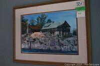 Framed signed print showing a lodge on rocky terrain with pine trees, glass reflection visible.