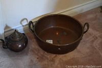 Photo showing the copper basin and kettle placed side by side on carpet, basin is large and round with handles, kettle has ornate detailing.