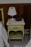 Painted wood night table with a central drawer and milk glass vase table lamp on top, with clock on table surface, showing overall condition and design.