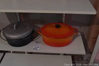 Side-by-side view of two Dutch ovens on a shelf: an orange/red Le Creuset enamelled Dutch oven and a grey McClary cast iron Dutch oven with lid.
