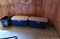 Three storage totes in unfinished room corner: two large blue Rubbermaid totes with beige lids closed, one smaller black tote on the left.