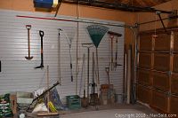 Wide view of garage wall with assorted gardening tools hanging and leaning against wall. Includes several rakes, shovels, pitchfork, triple claw weeder, seeder, and bags of seed. Bricks and pavers visible but not included.