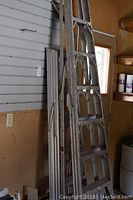 Two aluminum ladders standing upright against a wall inside a garage or workshop space. One ladder is shorter at about 6 feet with six steps, the other taller at about 8 feet with eight steps. Visible wear and tear marks show usage history.