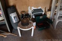 Photo showing green Coleman two-burner propane stove, small round portable charcoal or wood stove on a white plastic table, folded black and white camping chair, rolled tent bag, and some boxes.