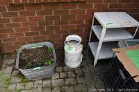 Overview photo of the metal wash tub filled with dirt, painted milk can beside it, and metal shelving unit to the right against a brick wall.