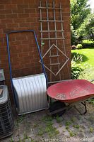 Wide shot of snow shovel with blue handle, wheelbarrow, and tall wooden trellis leaning against brick wall outdoors.