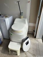 Front view of composting toilet with seat closed, vent stack pieces on top, boxed seat and step stool in front