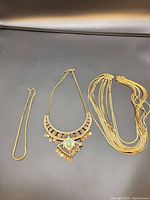 Three gold-tone necklaces laid out: curb chain, bib necklace with green stone, multi-strand necklace