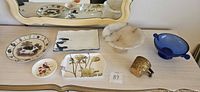 All eight items displayed on tabletop: pig plate, Asian plate, floral tray, bird dish, alabaster bowl, cobalt compote, brass cup, plate hanger visible